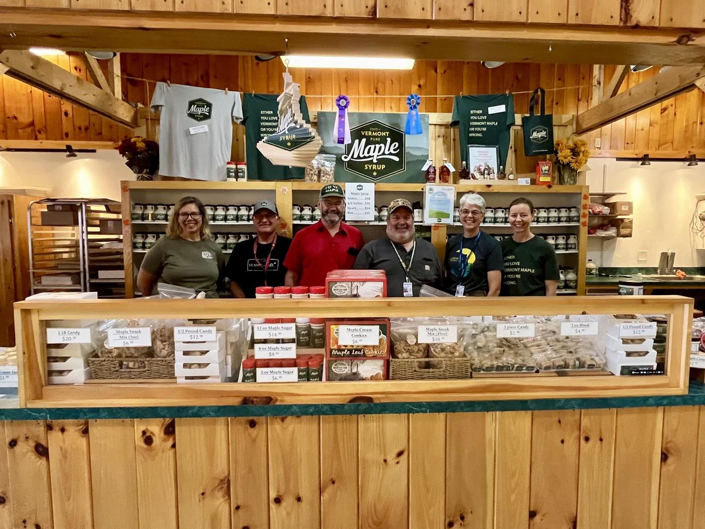 Farm stand with six smiling people behind a wooden counter with grain display.
