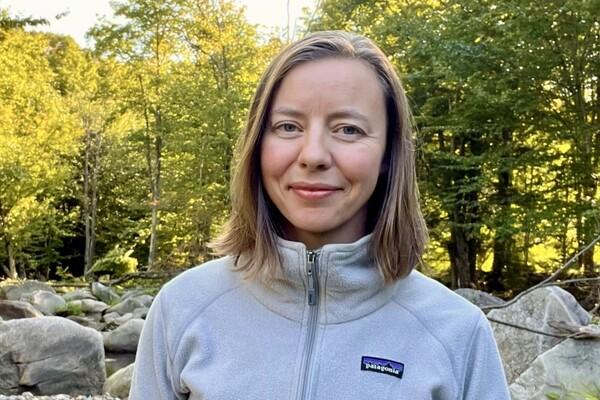 Woman in a light gray jacket smiling in a forest setting with rocks.
