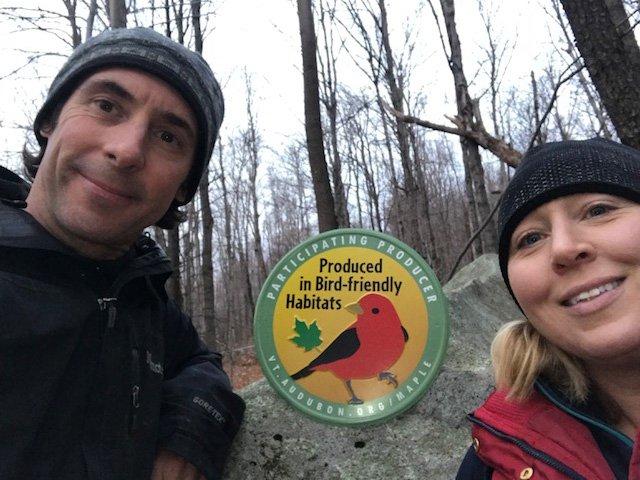 Two people in winter clothing beside a sign about bird-friendly habitats in a forest.