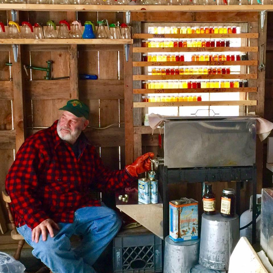 Man in a plaid shirt sits in a rustic cabin with bottles and jars on shelves.