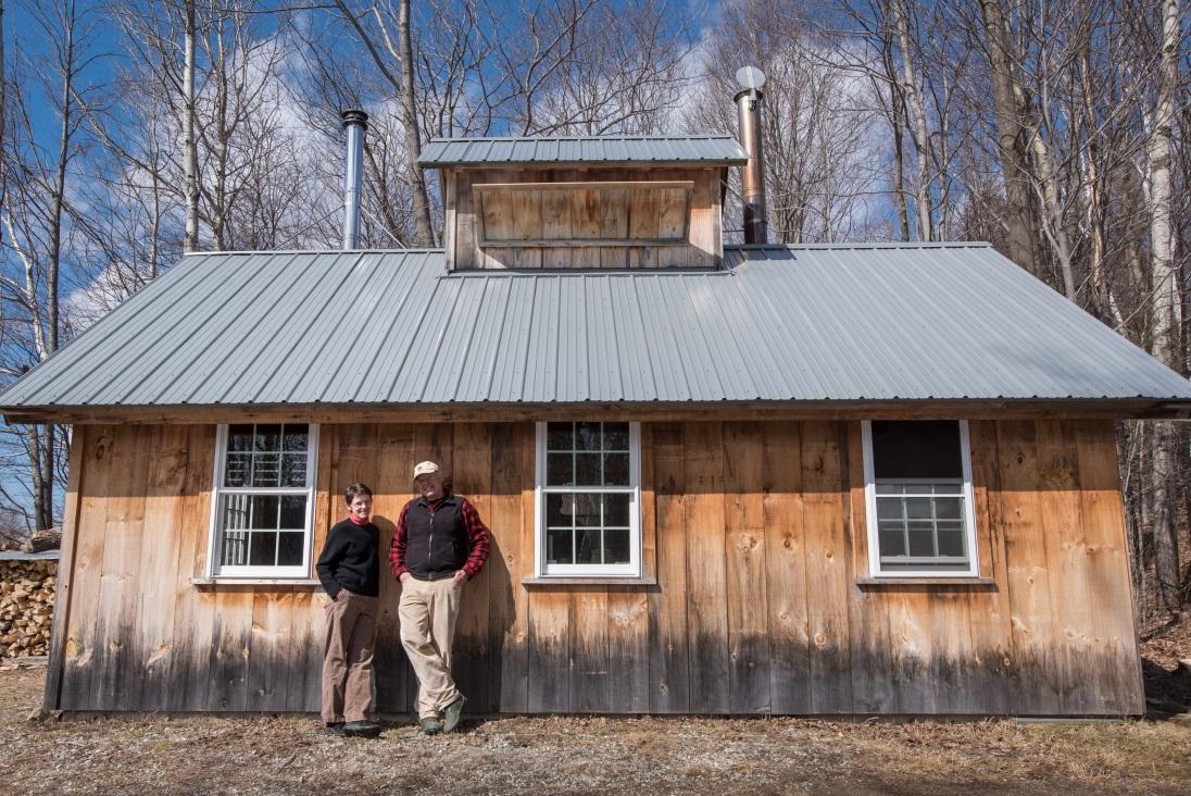 Two people lean against a wooden cabin with a metal roof, trees in the background.