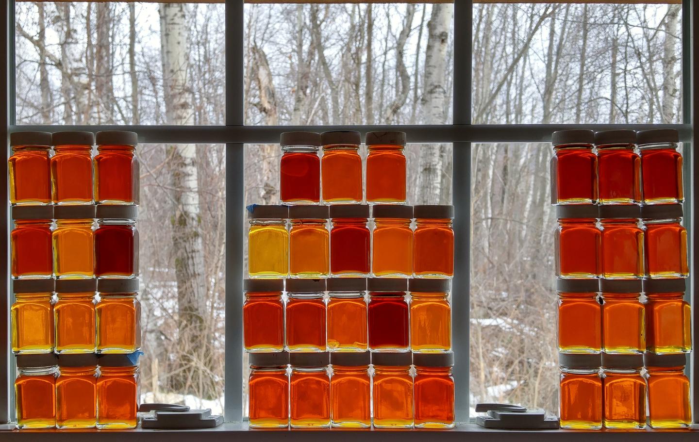 Jars of maple syrup in gradations of amber and black stacked in front of a snowy window view.
