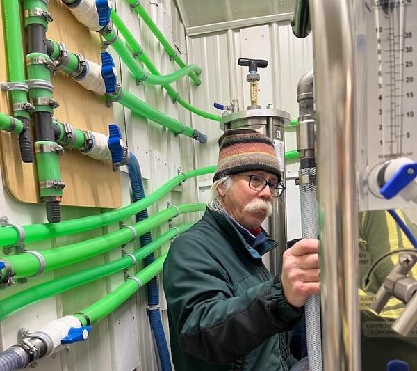 Man with glasses in a room, surrounded by green pipes and machinery.