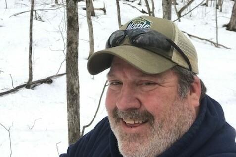 Smiling man in a snowy forest wearing a cap and sunglasses.
