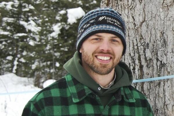 Man in winter clothing smiles outdoors by a tree in snowy woods.