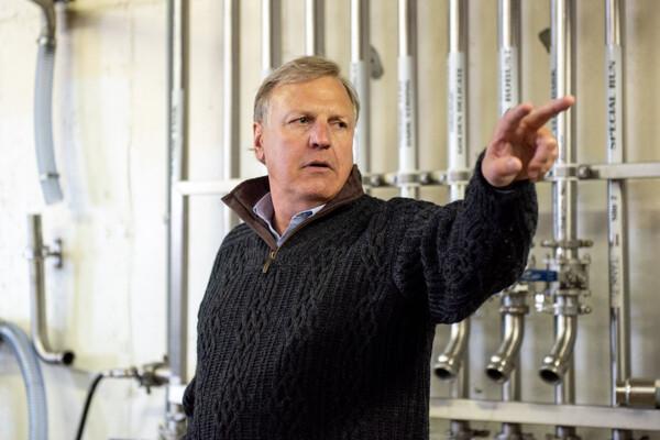 Man in a sweater gesturing in a room with pipes on the wall.