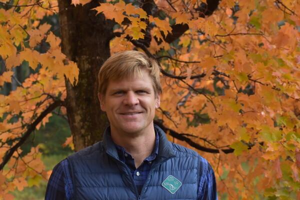 Man smiling in front of autumn trees with orange leaves.