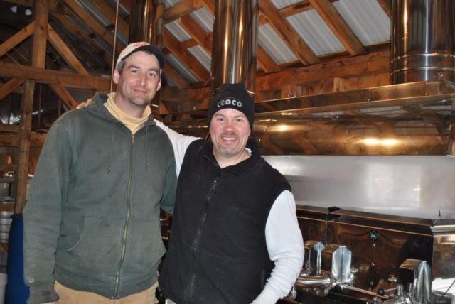 Two men smiling in a rustic wooden cabin, embracing each other.