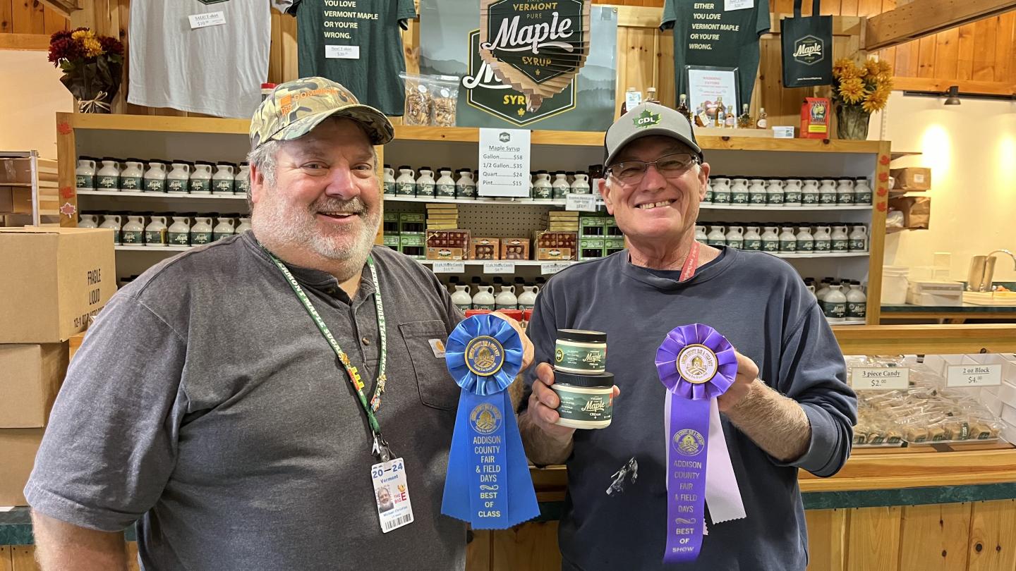 Two men smiling, holding award ribbons and jars in a market setting.
