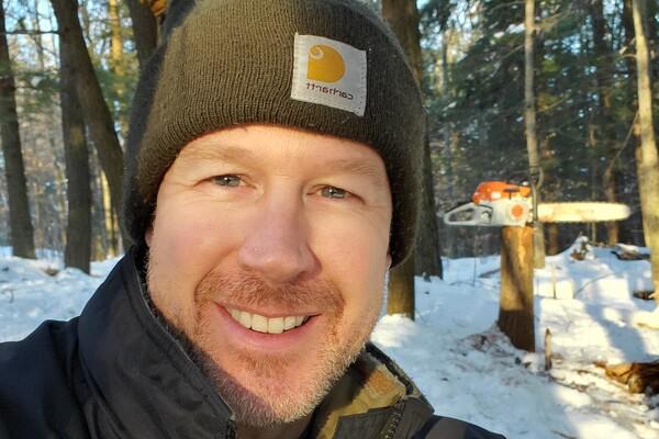 Man smiling outdoors in snowy forest, wearing a beanie and jacket.