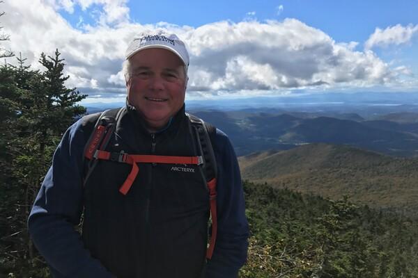 Smiling man on a mountain with a cloudy sky and distant peaks.