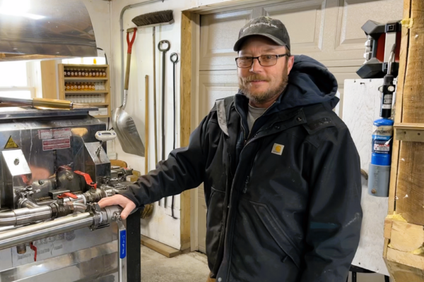 Man in a workshop, wearing a black jacket and cap, stands beside machinery.