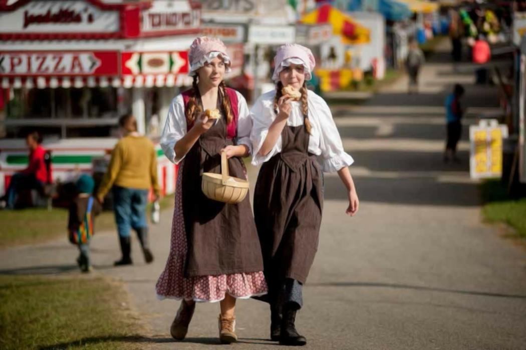 Two women in old-fashioned dresses walk at a fair holding ice cream cones.