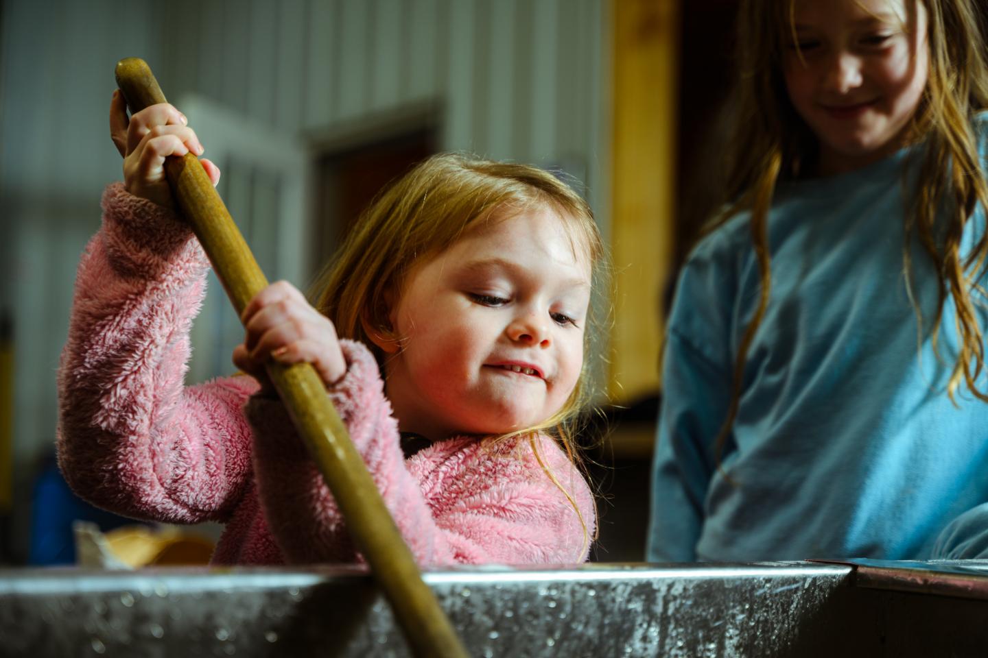Young child stirring a pot with a stick, smiling adult in a blue sweater nearby.