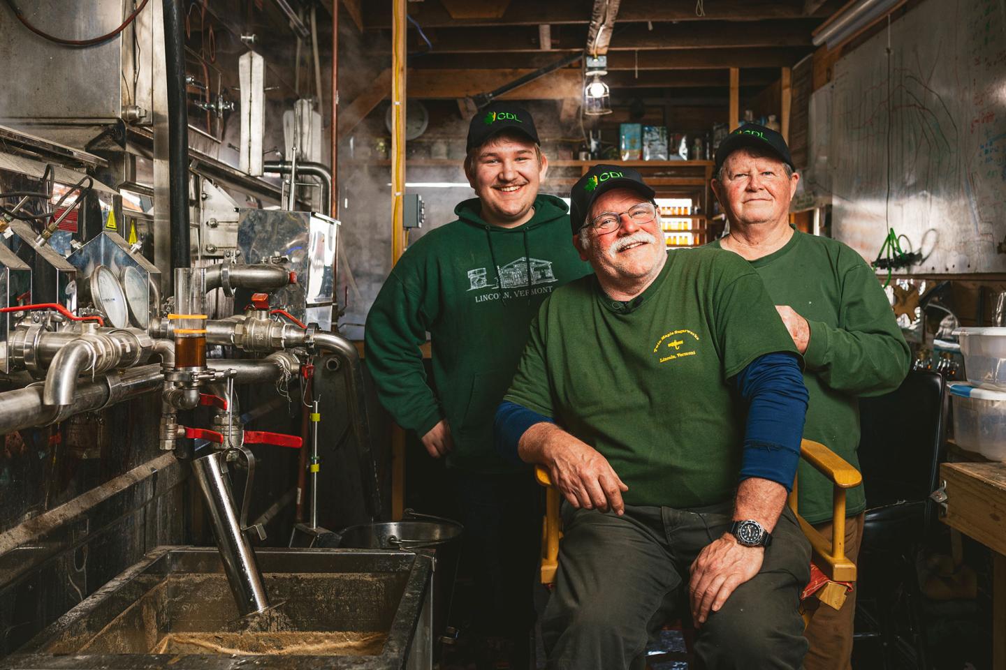 Three people in green shirts and hats smiling in an industrial setting.