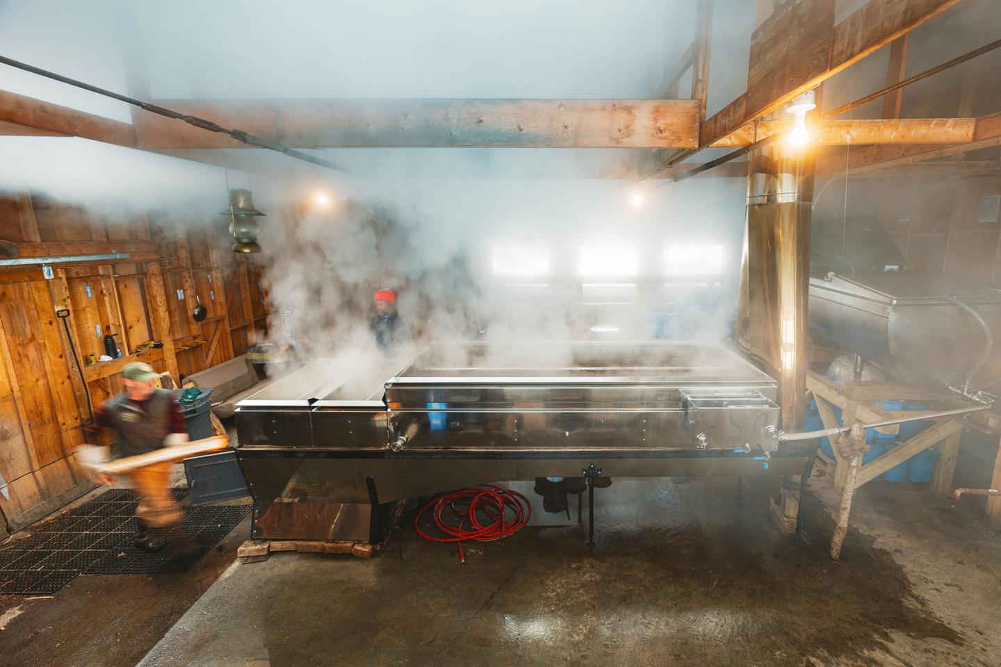 Rustic kitchen with large metal cooking vats steaming, person moving quickly in foreground.