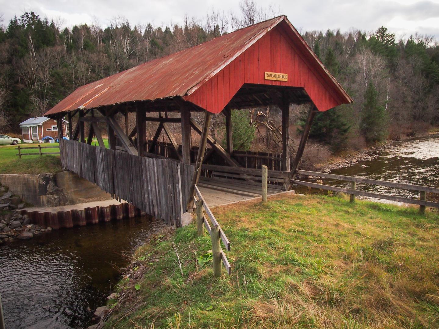 Red-roofed wooden covered bridge over a small creek, surrounded by trees and grass.