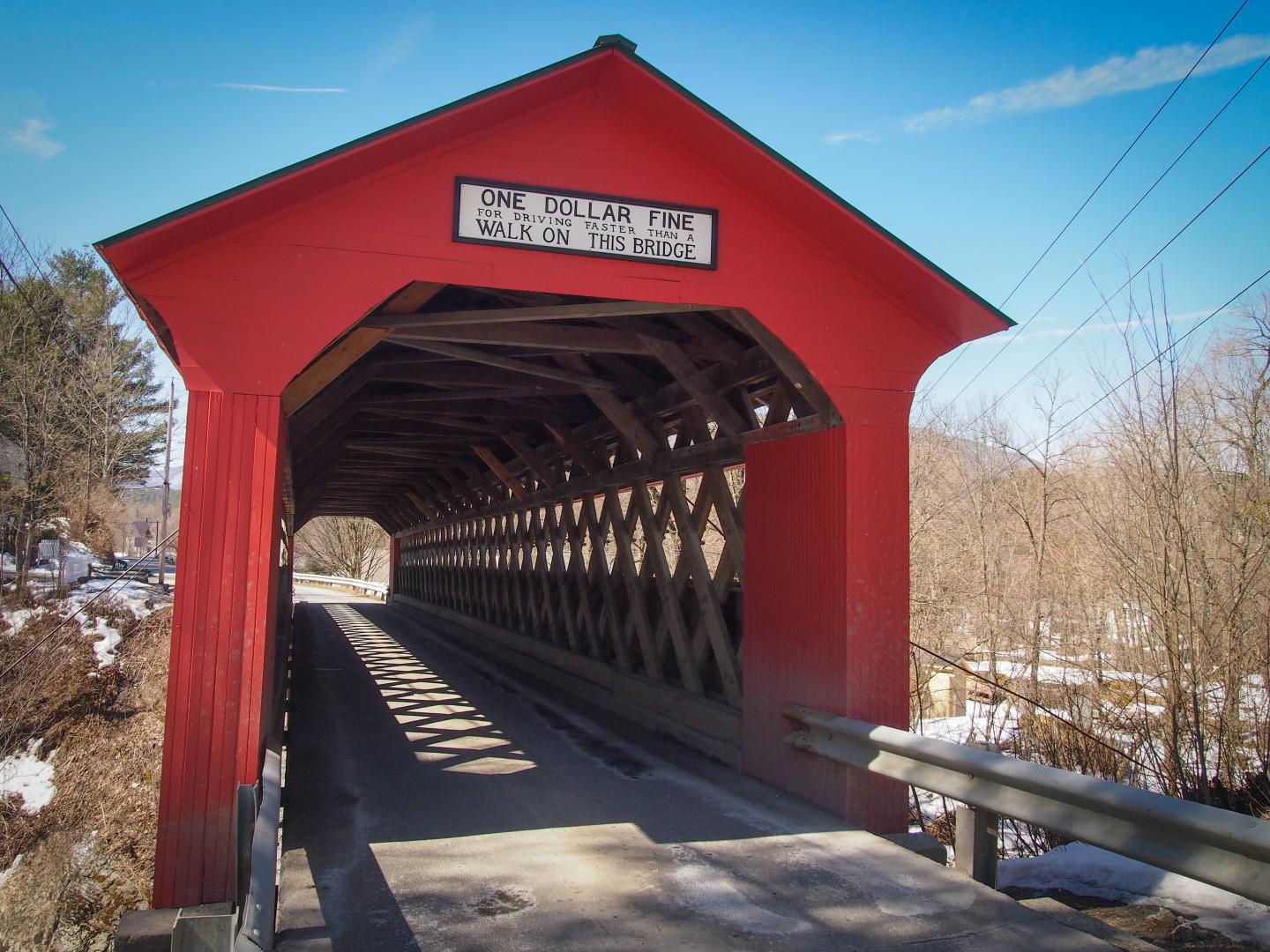 Red covered bridge with blue sky, surrounded by bare trees.