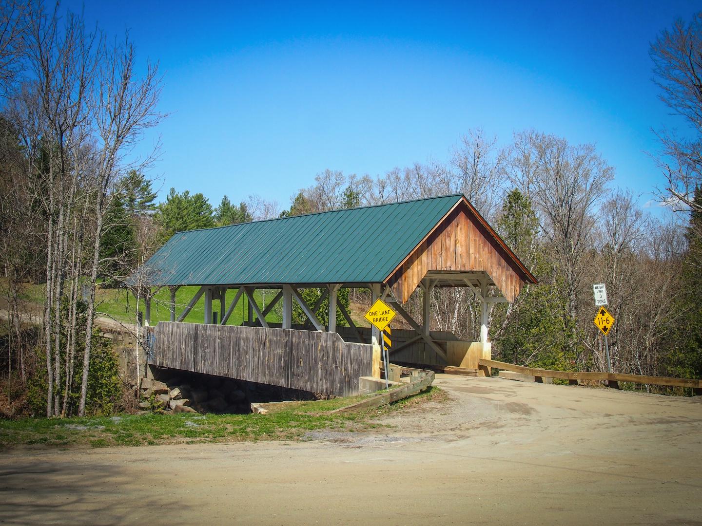Covered bridge in a rural setting with forested background under clear blue sky.