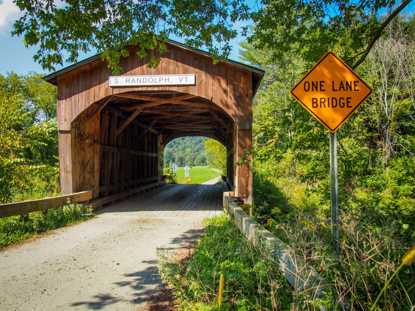 Wooden covered bridge with a "One Lane Bridge" sign amidst lush greenery.