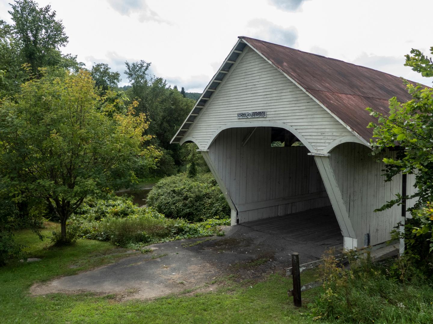 Old covered bridge with rusted roof surrounded by trees and greenery.