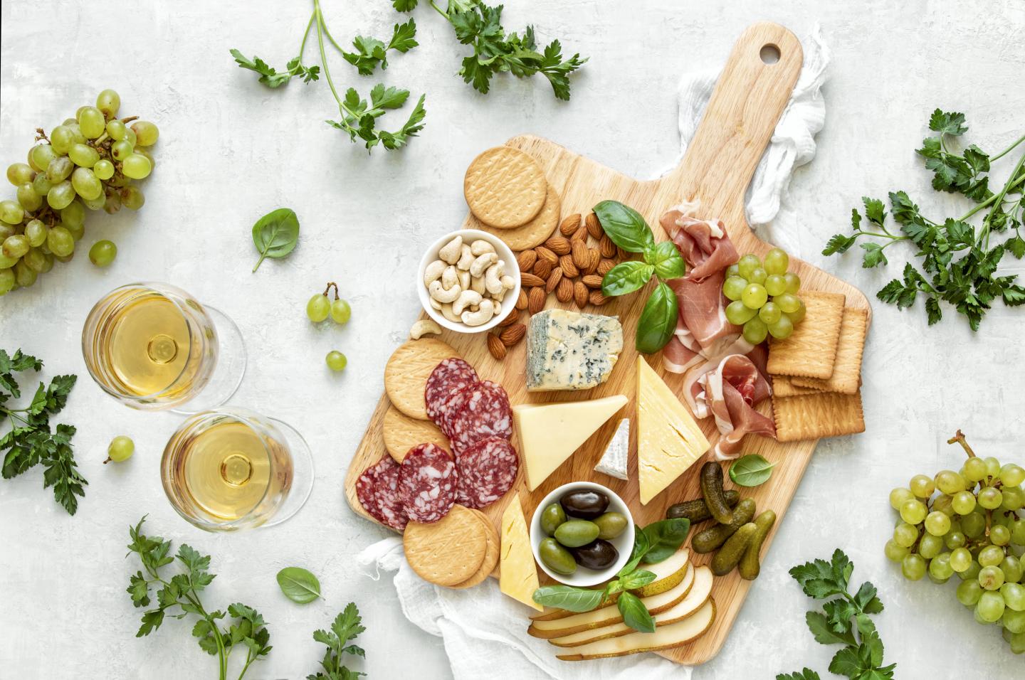 Charcuterie board with cheeses, meats, crackers, and olives, surrounded by wine glasses and grapes.