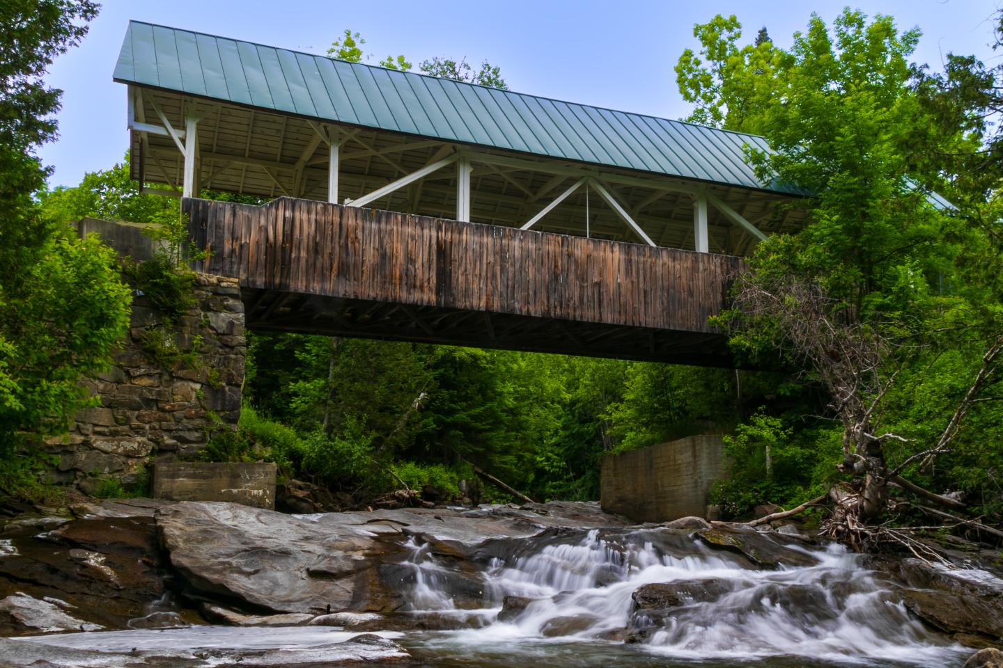 Covered bridge over a rocky stream surrounded by green trees.