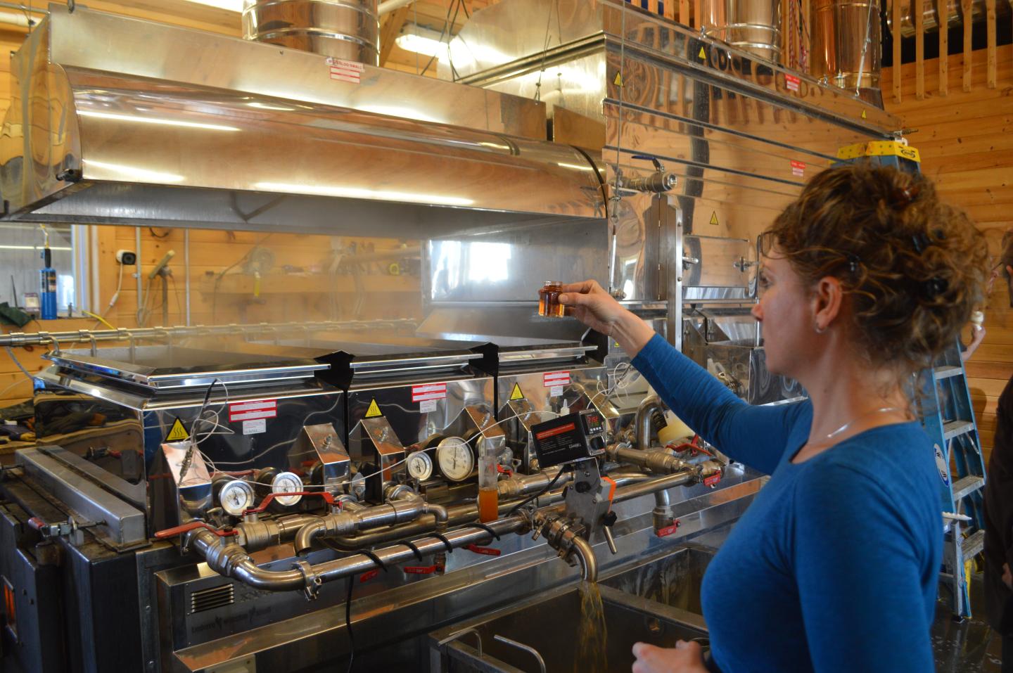 Woman operating syrup production equipment indoors, wearing blue shirt.
