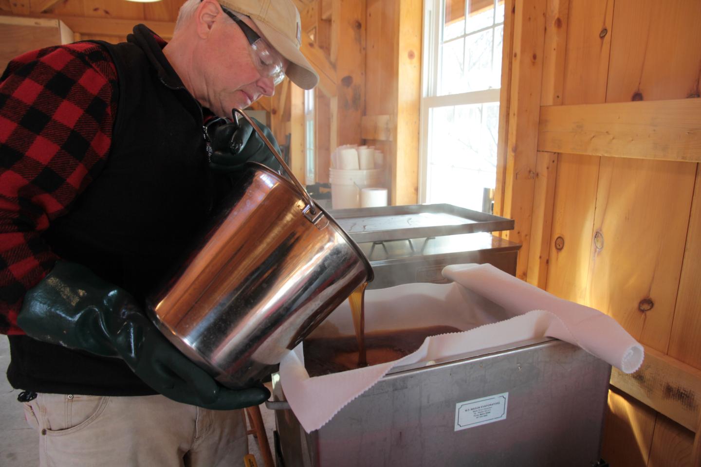 Man pouring syrup into a container inside a wooden cabin.