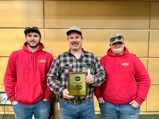 Three men wearing caps, middle holding a plaque, in front of a wooden wall.