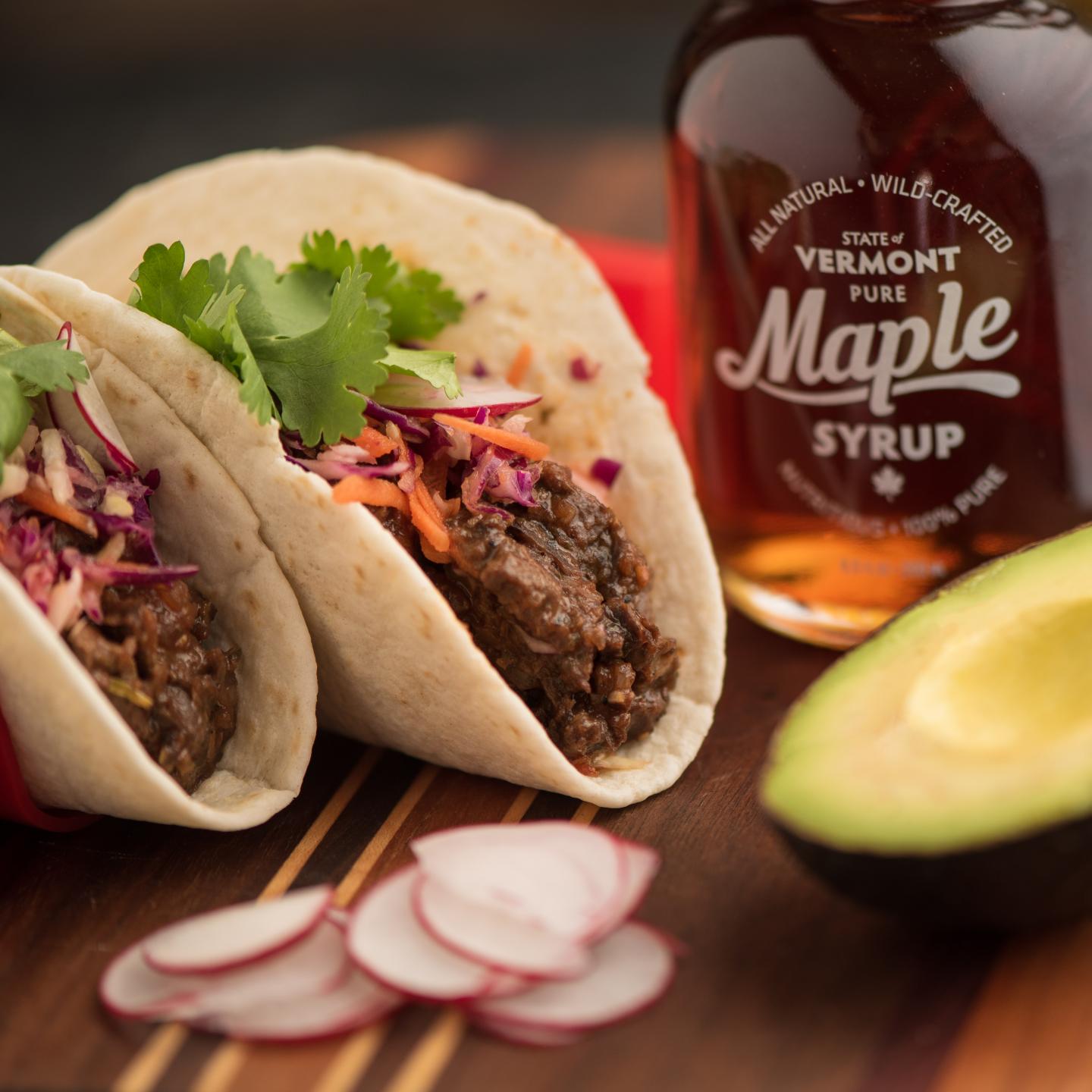 Two tacos with cilantro and slaw, maple syrup bottle, sliced radishes, and avocado.