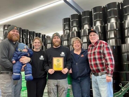 Group smiling, one holding a plaque, surrounded by stacked barrels indoors.