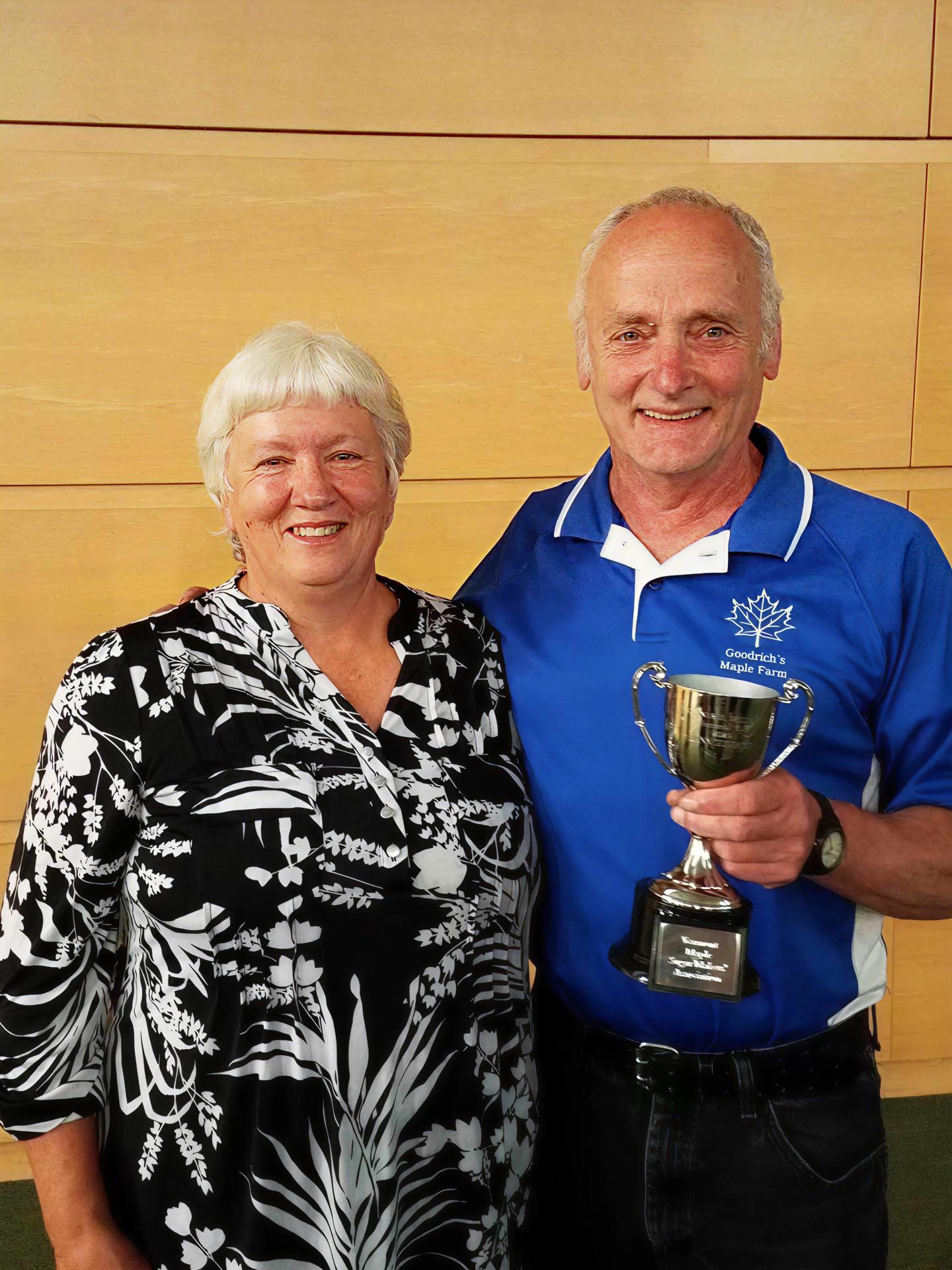 Man and woman smiling, man holding trophy, against wooden wall.