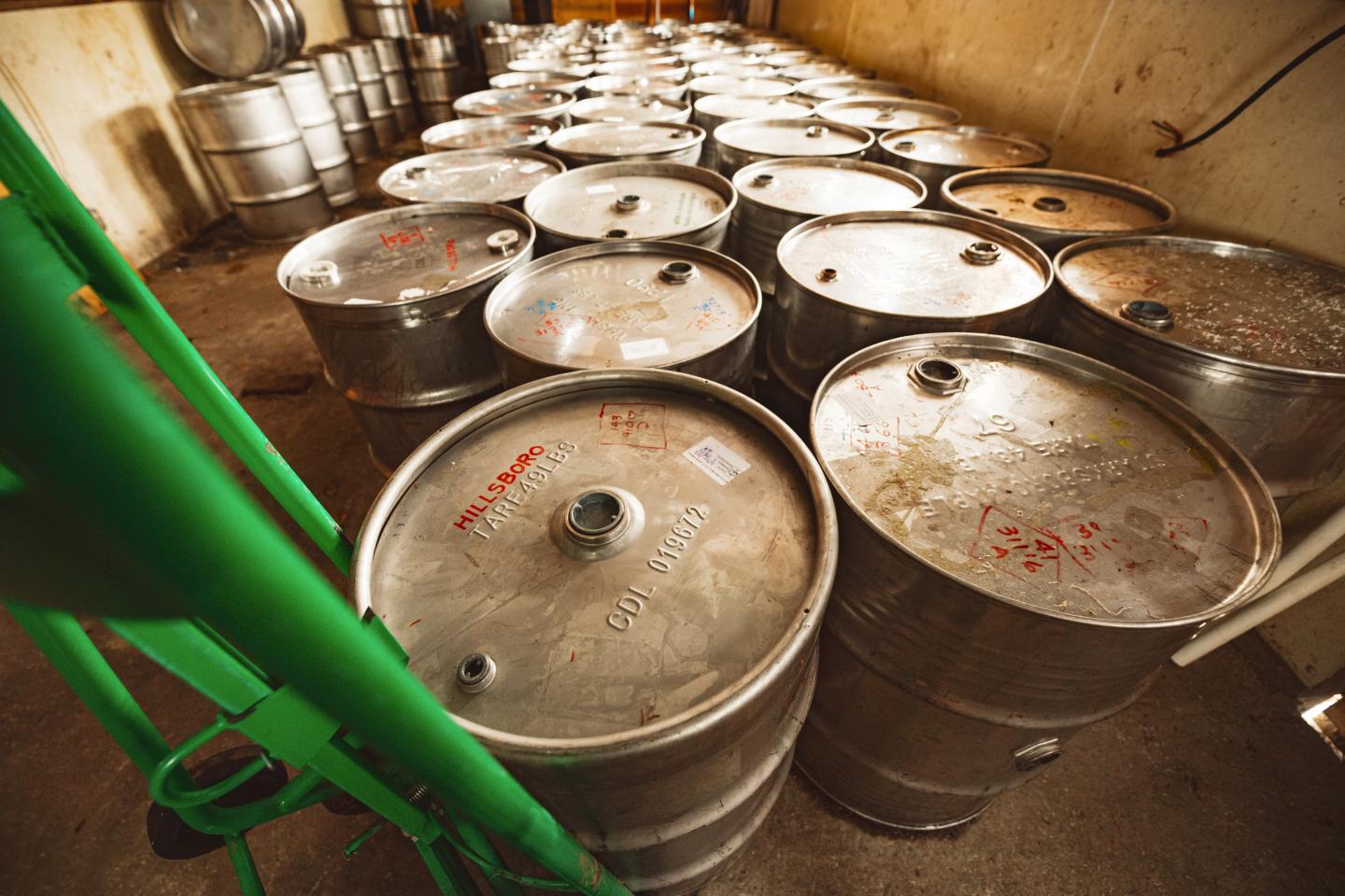 Storage room with rows of metal beer kegs, some marked with text, and a green hand truck.