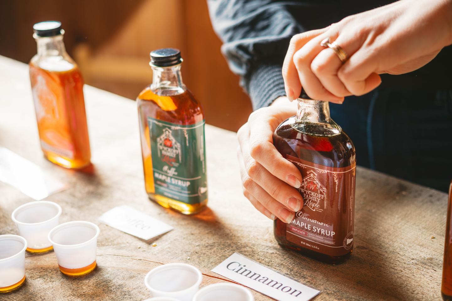 Hands opening a bottle of maple syrup on a wooden table.