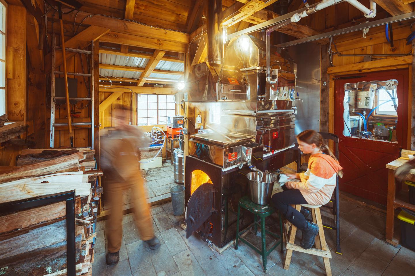 Rustic wooden workshop with a person moving and another seated near a stove. Warm lighting.