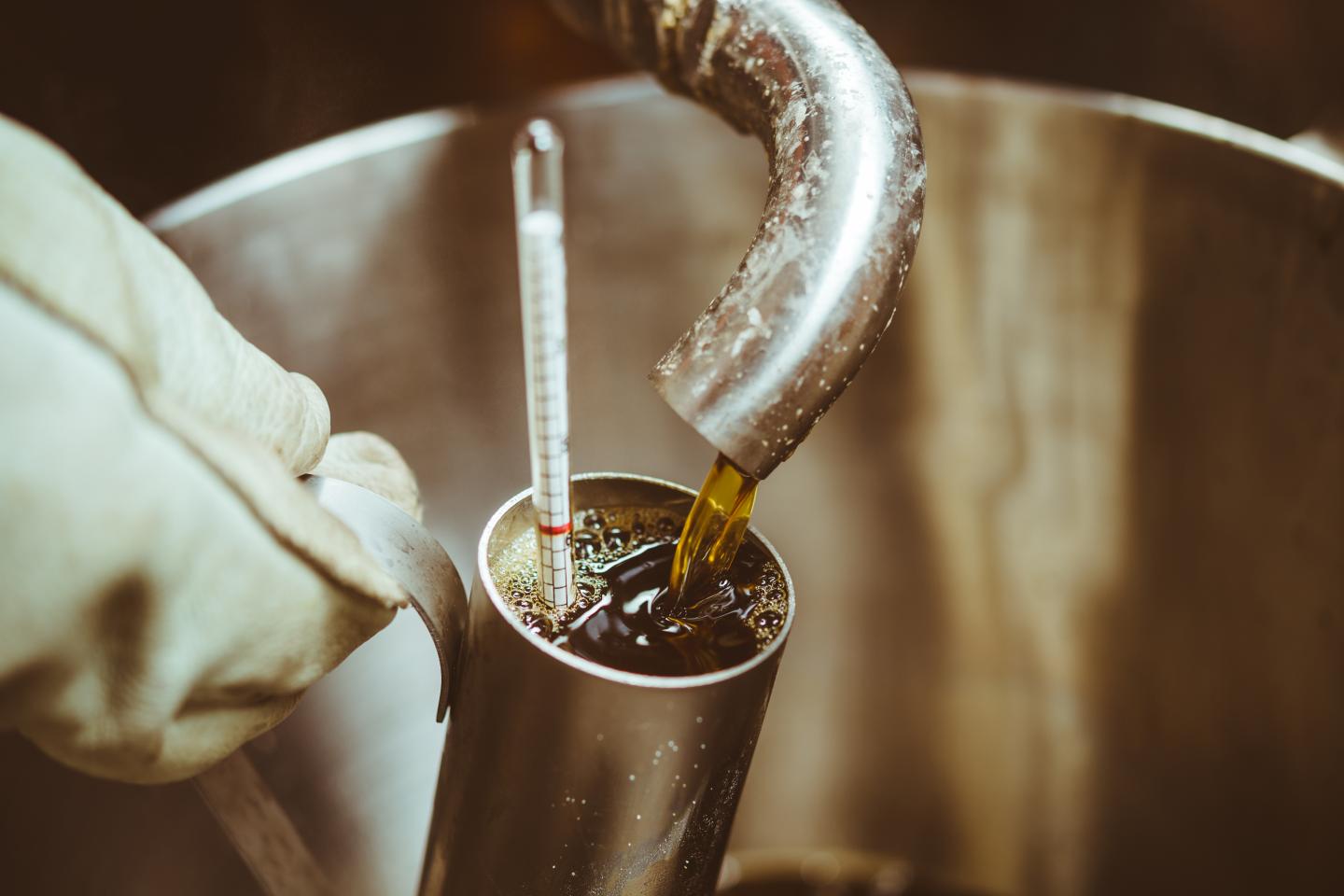 Metal tube being filled with syrup under a curved pipe, thermometer inside.