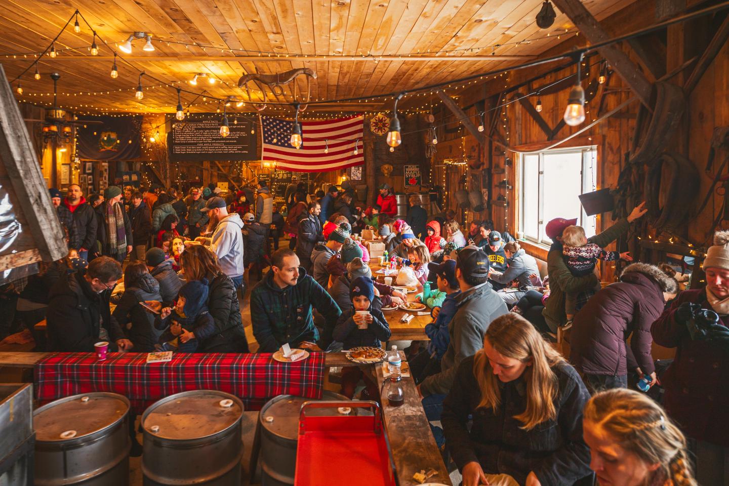 Rustic barn gathering with people dining under warm lights, U.S. flag in the background.