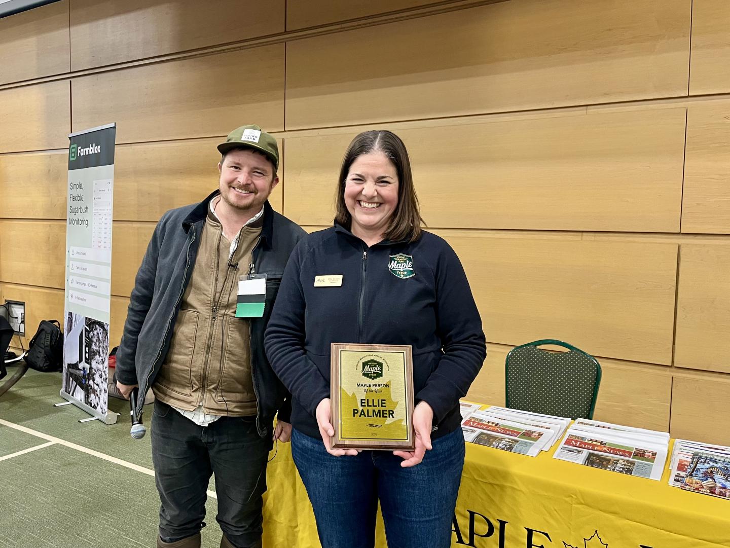 Two people smiling, one holding a plaque, indoors at an event.