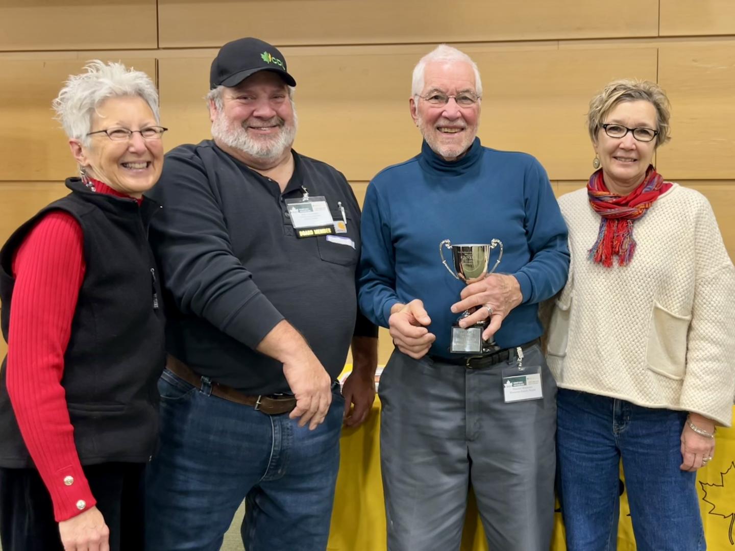 Four smiling adults, one holding a trophy, standing together indoors.