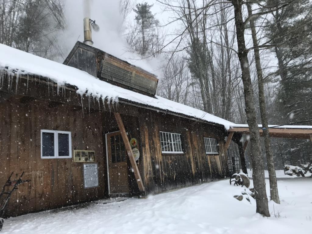 Rustic wooden sugarhouse with icicles, surrounded by snow and trees.