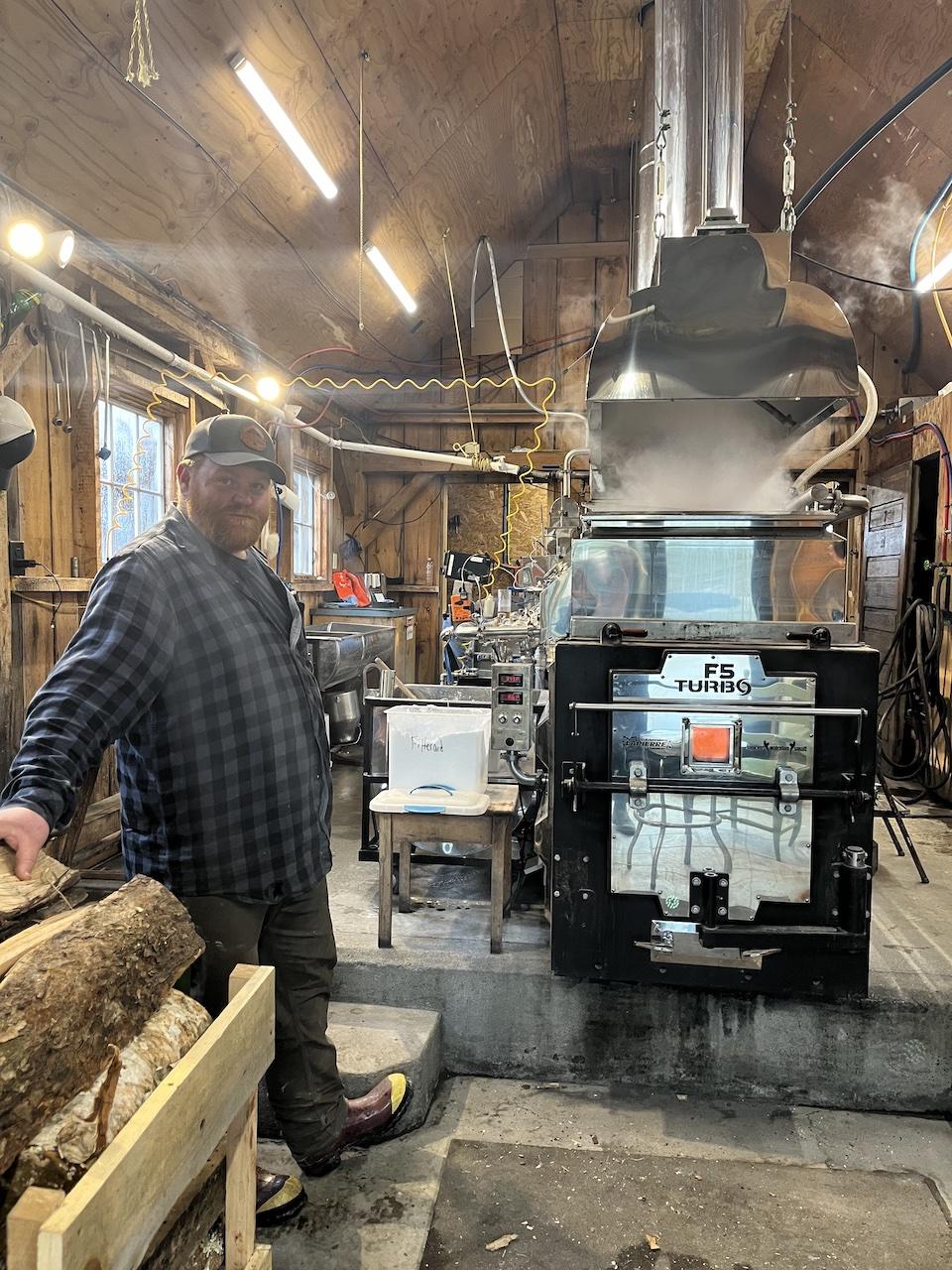 Man in a flannel shirt stands by a large metal stove in a wooden cabin.