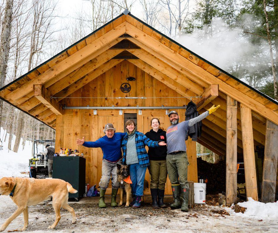 Four people and a dog stand happily in front of a wooden cabin in a snowy forest.