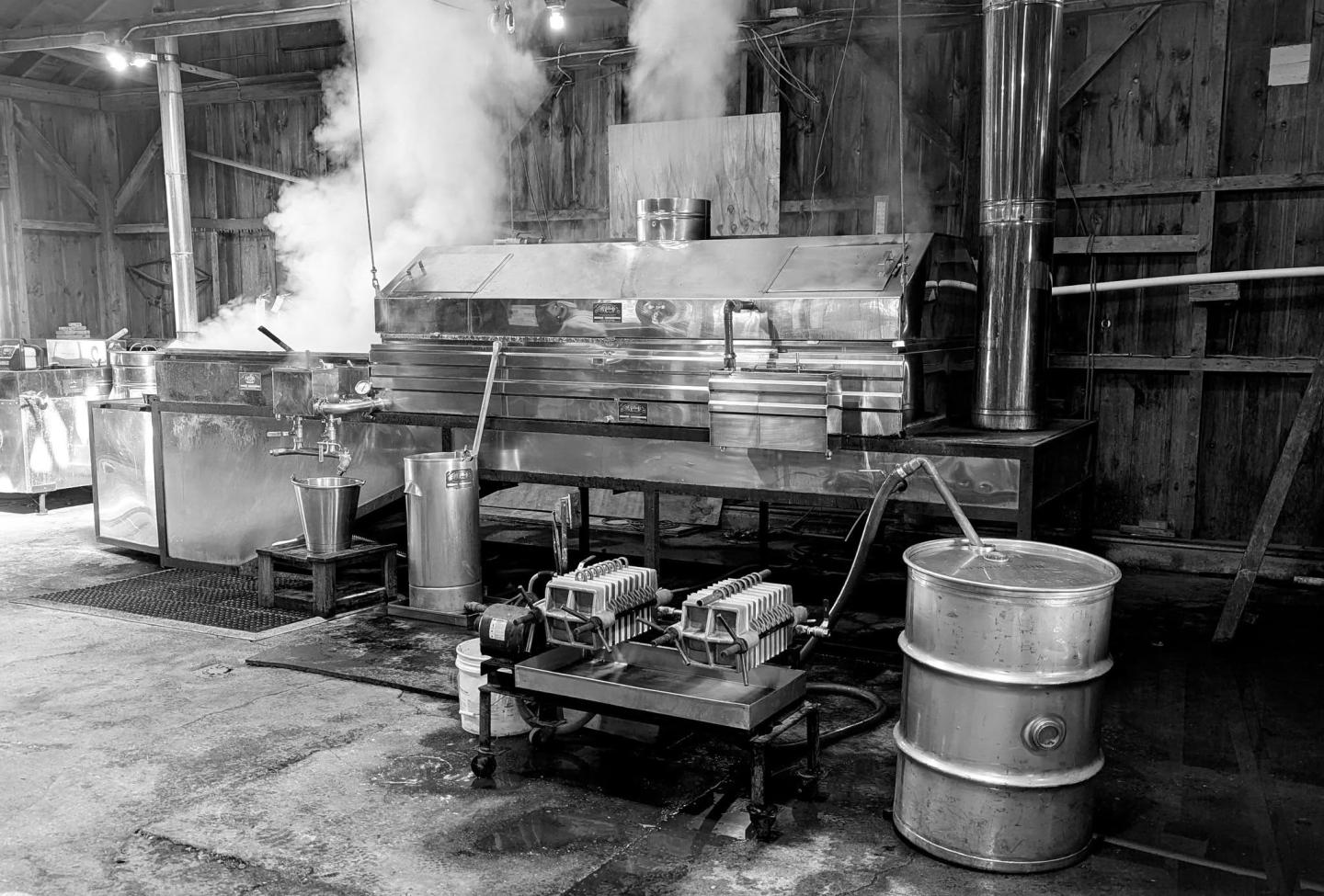 Black and white photo of a syrup evaporator in a rustic wooden building.