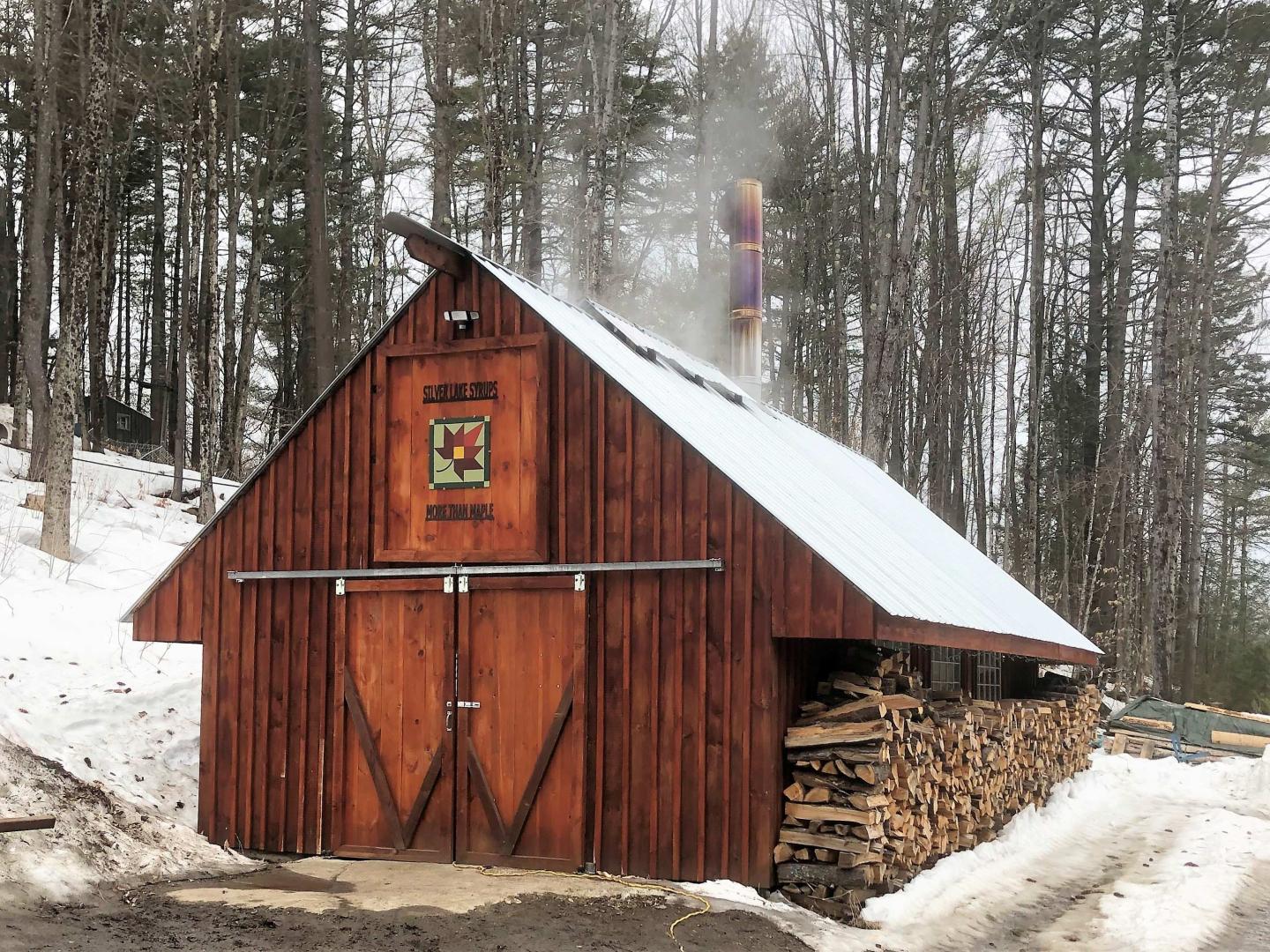 Wooden sugar shack with smoke rising, snow and stacked firewood nearby.