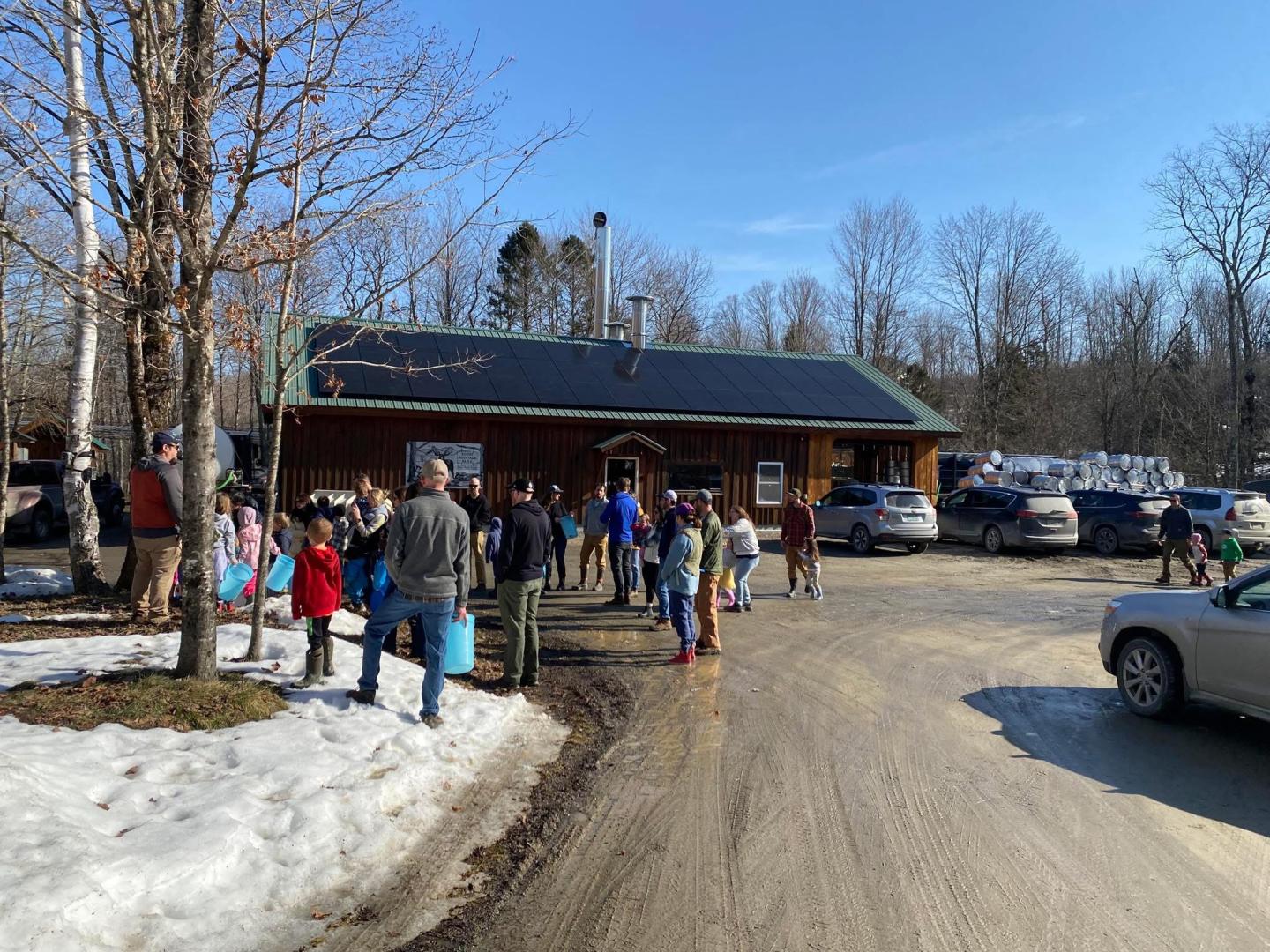 Group of people outside a wooden building on a sunny day with snow on the ground.