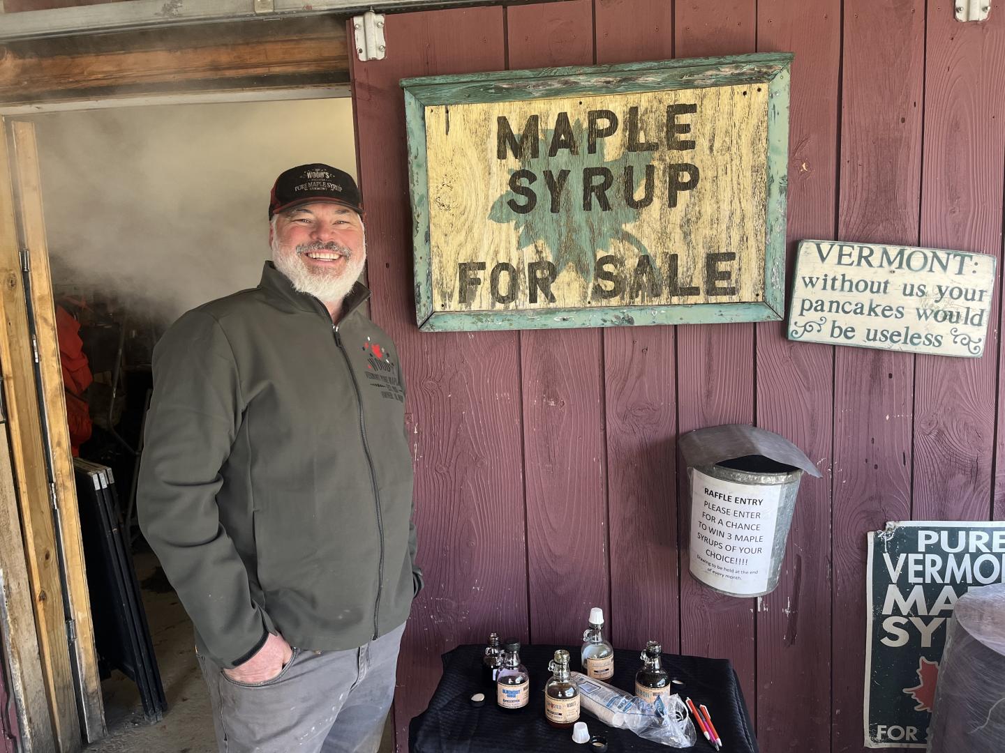 Smiling man stands by a "Maple Syrup for Sale" sign on a rustic wall, with maple syrup bottles.