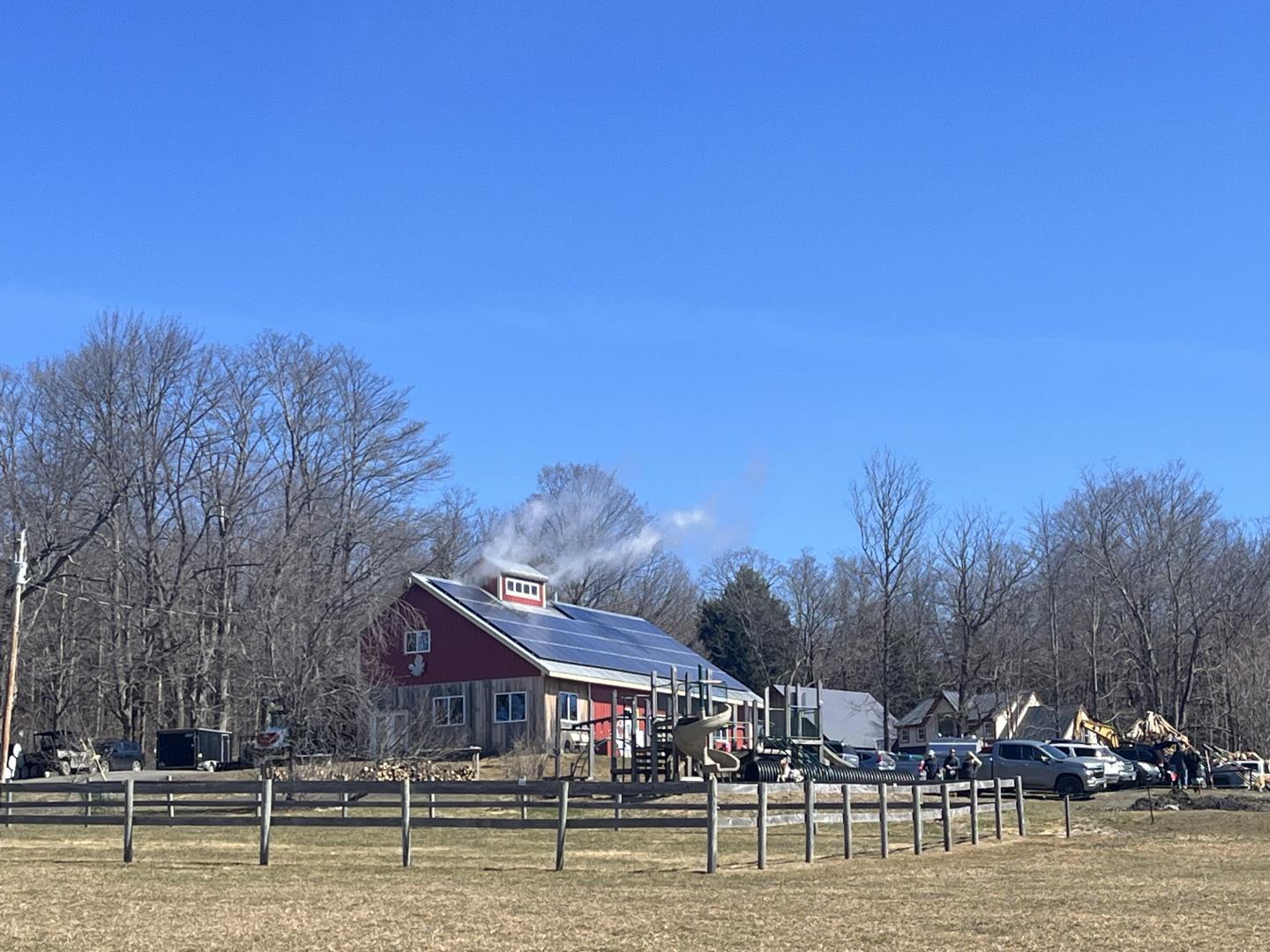 Red barn with solar panels on the roof, surrounded by bare trees and a clear blue sky.