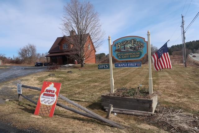 Country store sign with U.S. flag, red house in the background, clear blue sky.