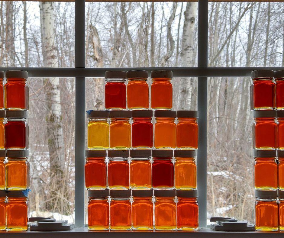 Jars of maple syrup in warm hues on a windowsill, snowy trees outside.